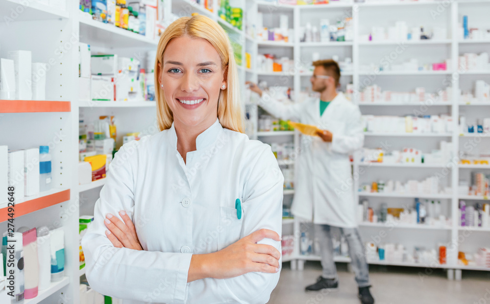 Portrait of a female pharmacist, male colleague working with drugs in the background.