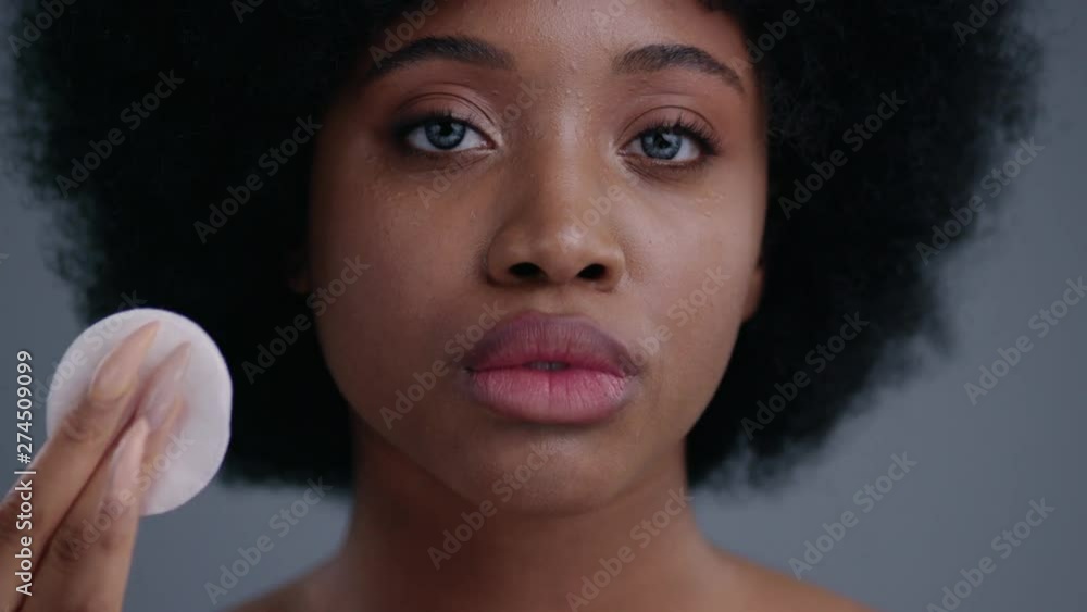 Close-up beautiful portrait of african young woman cleaning her tender face with a cotton disc. Amazing black girl smiling on camera on grey background. Facial skin care routine. Close-up beautiful portrait of african young woman cleaning her tender face with a cotton disc. Amazing black girl smiling on camera on grey background. Facial skin care routine.