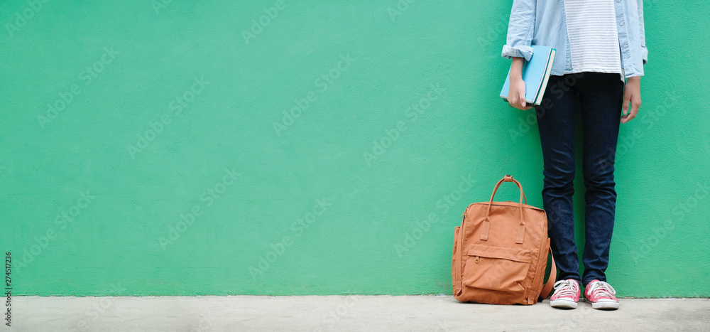 Student girl holding books and school bag standing over green wall background with copy space, education banner, back to school concept - obrazy, fototapety, plakaty 