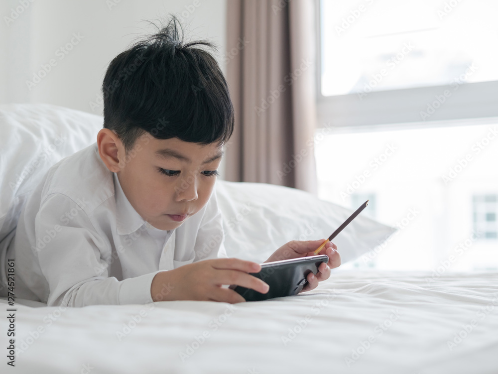 Cute little asian kid focused on smartphone while lying on the bed. Boy playing games on smartphone. Social and technology concept Cute little asian kid focused on smartphone while lying on the bed. Boy playing games on smartphone. Social and technology concept