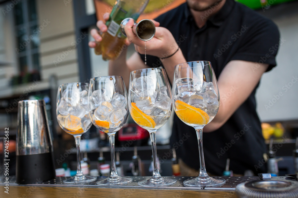 four glasses standing on the bar. Glasses are with ice and orange. Barman pours alcohol in glasses from a measuring glass four glasses standing on the bar. Glasses are with ice and orange. Barman pours alcohol in glasses from a measuring glass