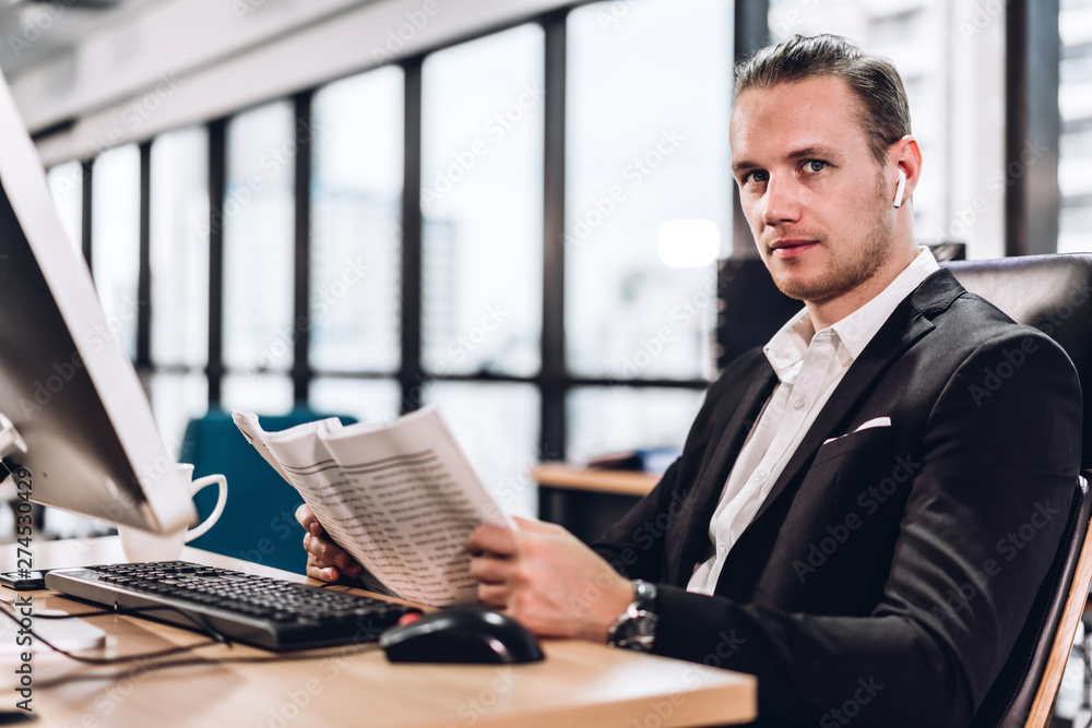 Confident young handsome businessman reading newspaper and working with computer in modern office Confident young handsome businessman reading newspaper and working with computer in modern office