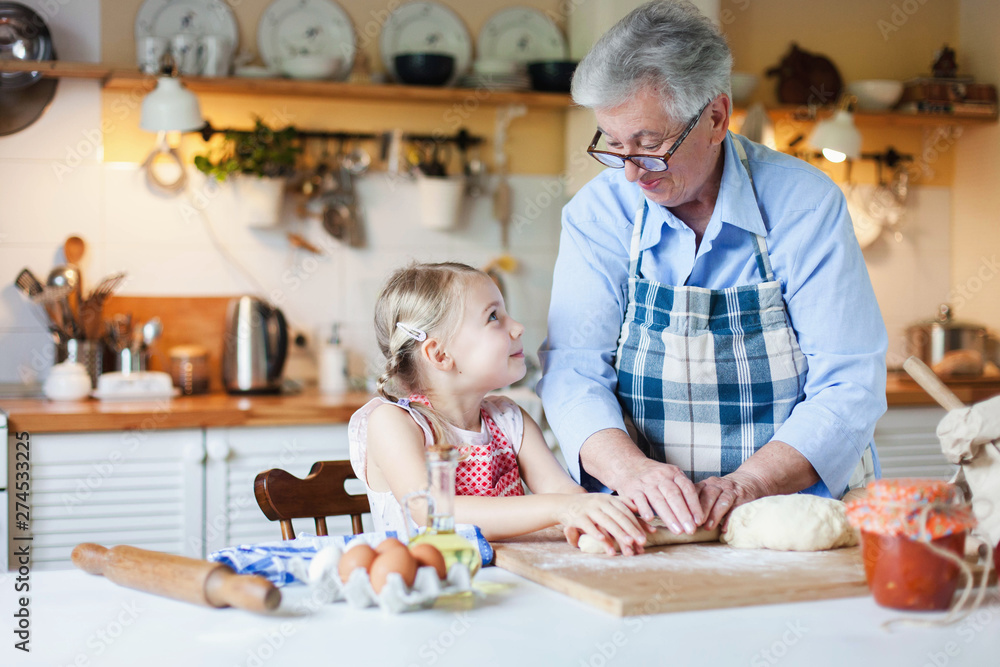 Fototapeta premium Family is cooking in cozy home kitchen. Grandmother and granddaughter are happy together. Senior woman and little child girl are smiling. Cute kid is helping to prepare for Thanksgiving Day dinner.