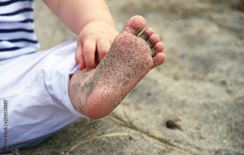 Baby girl wearing white pants and striped shirt, sitting in sand at Kitsilano Beach, Vancouver BC, Canada, showing her sandy foot.