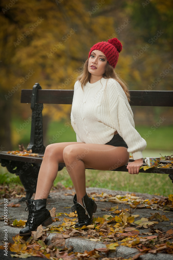 Portrait of a beautiful young Caucasian sensual woman with a red cap ,white pullover and black boots in autumn park, red green yellow threes. Girl with long legs and a cup of coffee siting on bench