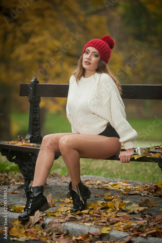 Obraz na płótnie Portrait of a beautiful young Caucasian sensual woman with a red cap ,white pullover and black boots in autumn park, red green yellow threes. Girl with long legs and a cup of coffee siting on bench w salonie