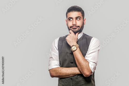 Portrait of thoughtful or confused handsome bearded brunette man in white shirt and waistcoat standing, holding his chin, looking away and thinking. indoor studio shot isolated on gray background.