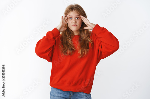 Girl shocked cannot understant what happening standing confused and perplexed with mind blowing out from information holding hands on temples lifting eyebrows questioned wearing red sweater