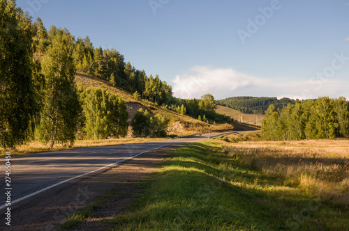 Asphalt road going across mountains and green forests. Trees and their shadows on the grass. Sunny summer day with blue sky. Ural landscape