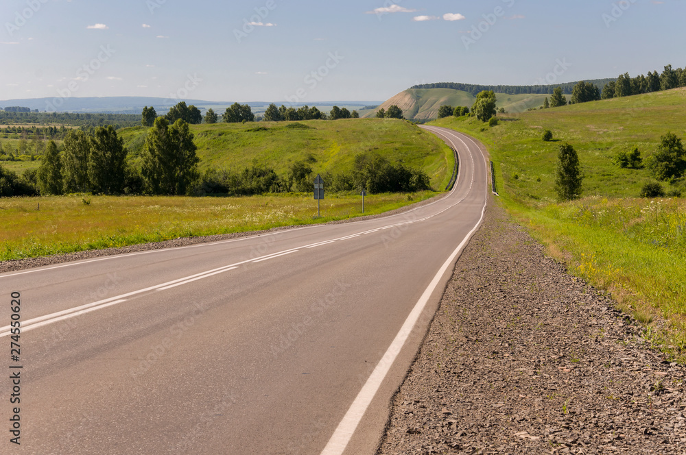 Asphalt road going across mountains and green forests. Trees and their shadows on the grass. Sunny summer day with blue sky. Ural landscape