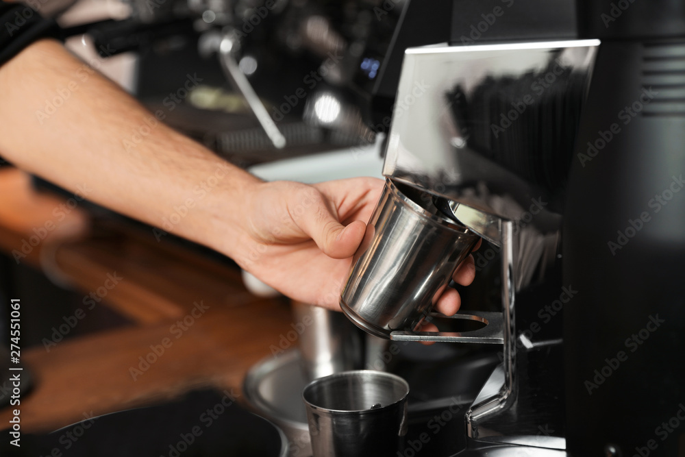 Barista with metal cup using coffee grinding machine in cafe, closeup