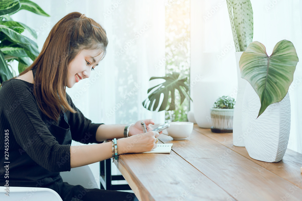Portrait of young beautiful woman using smartphone to working or shopping online and writing on notebook in cafe with cup of coffee on wooden table Portrait of young beautiful woman using smartphone to working or shopping online and writing on notebook in cafe with cup of coffee on wooden table