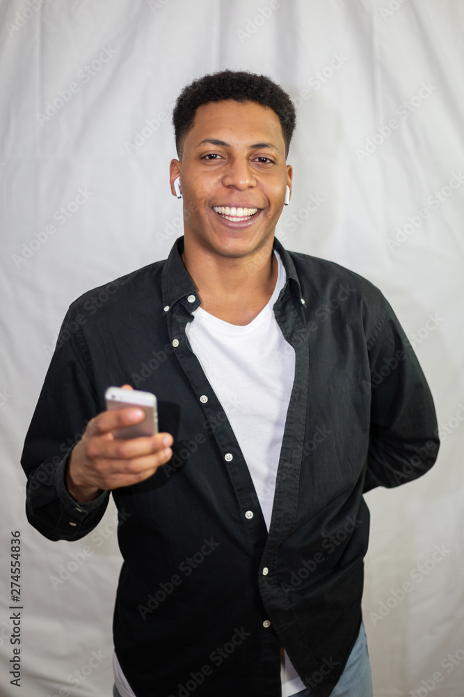 Happy African American young handsome man with wireless earphones in black shirt holding smartphone and laughing Happy African American young handsome man with wireless earphones in black shirt holding smartphone and laughing