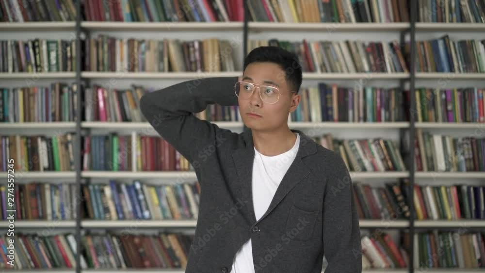 Gimbal middle shot of asian man in eyeglasses choosing a book from bookshelf scratching his head. Asian man pointing at a book on background of bookshelves in library Gimbal middle shot of asian man in eyeglasses choosing a book from bookshelf scratching his head. Asian man pointing at a book on background of bookshelves in library
