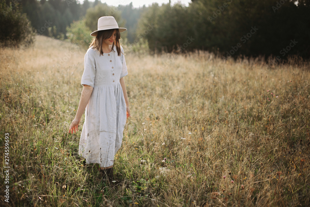 Stylish girl in linen dress walking among herbs and wildflowers in sunny meadow in mountains. Boho woman relaxing in countryside, simple rustic life style. Atmospheric image. Space text Stylish girl in linen dress walking among herbs and wildflowers in sunny meadow in mountains. Boho woman relaxing in countryside, simple rustic life style. Atmospheric image. Space text