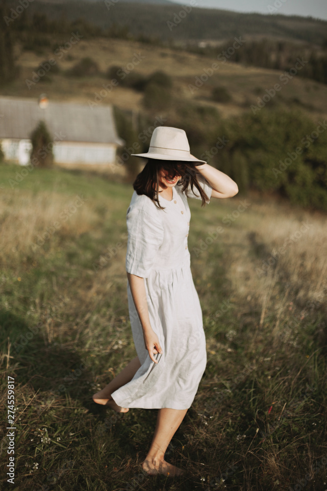 Stylish girl in linen dress and hat running barefoot in grass in sunny field at village. Boho woman relaxing in countryside, simple rustic life. Atmospheric image. Space text