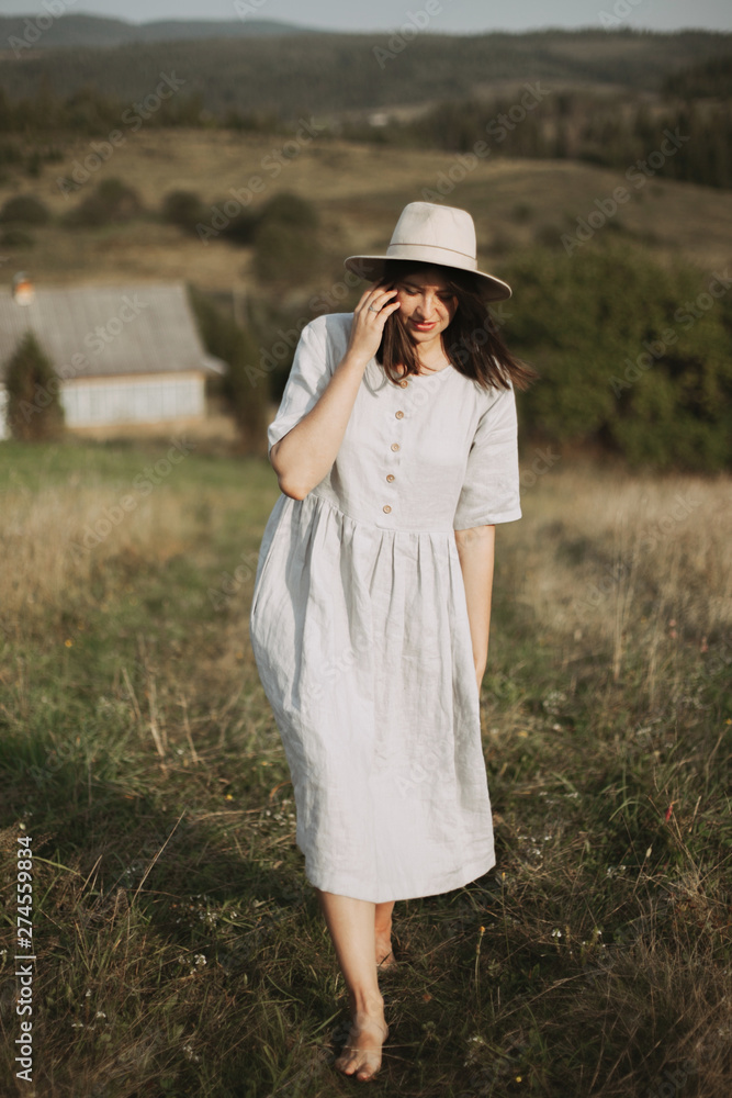 Stylish girl in linen dress and hat walking barefoot in grass in sunny field at village. Boho woman relaxing in countryside, simple rustic life. Atmospheric image. Space text