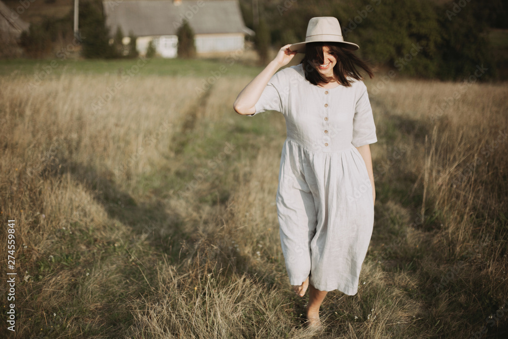 Obraz premium Stylish girl in linen dress and hat walking barefoot among herbs and wildflowers in sunny field in mountains. Boho woman smiling in countryside, simple rustic life. Atmospheric image