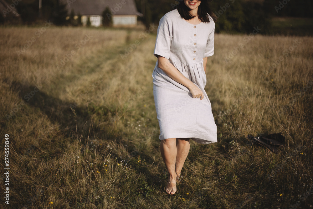 Stylish girl in linen dress and hat running barefoot in grass in sunny field at village. Boho woman relaxing in countryside, simple rustic life. Atmospheric image. Space text Stylish girl in linen dress and hat running barefoot in grass in sunny field at village. Boho woman relaxing in countryside, simple rustic life. Atmospheric image. Space text