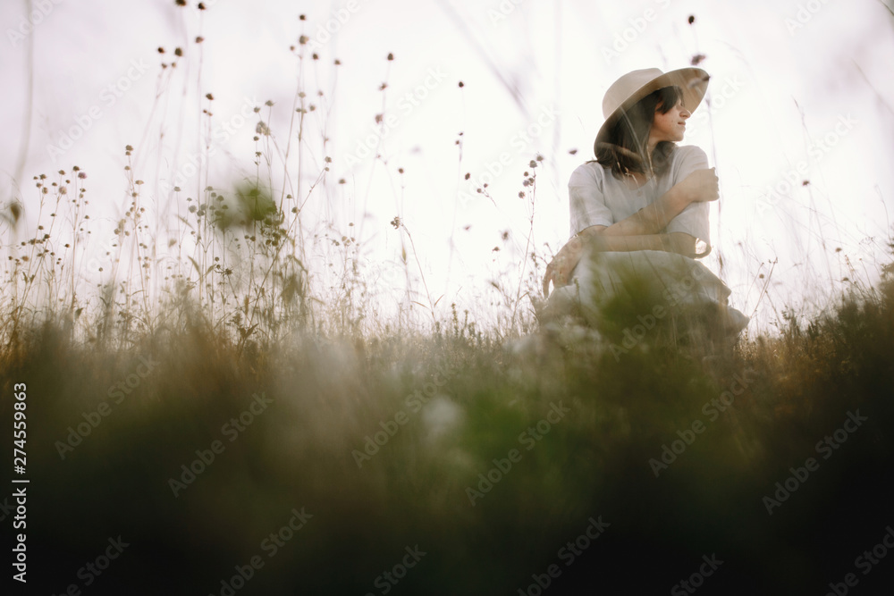 Obraz premium Stylish girl in rustic dress and hat sitting among wildflowers and herbs in sunny meadow in mountains. Boho woman relaxing in countryside, simple life. Atmospheric image. Space text