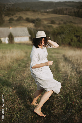Stylish girl in linen dress running barefoot among herbs and wildflowers in sunny field in mountains. Boho woman relaxing in countryside, simple rustic life. Atmospheric image. Space text © sonyachny