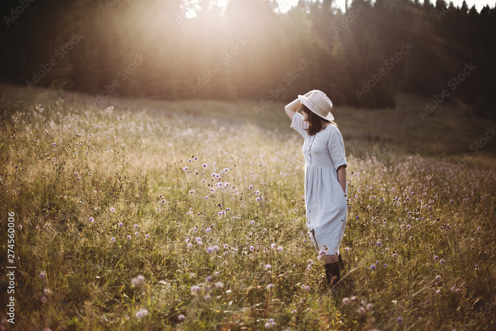 Stylish girl in rustic dress walking in wildflowers in sunny meadow in mountains. Boho woman relaxing in countryside flowers at sunset, rural simple life. Atmospheric image. Space text Stylish girl in rustic dress walking in wildflowers in sunny meadow in mountains. Boho woman relaxing in countryside flowers at sunset, rural simple life. Atmospheric image. Space text