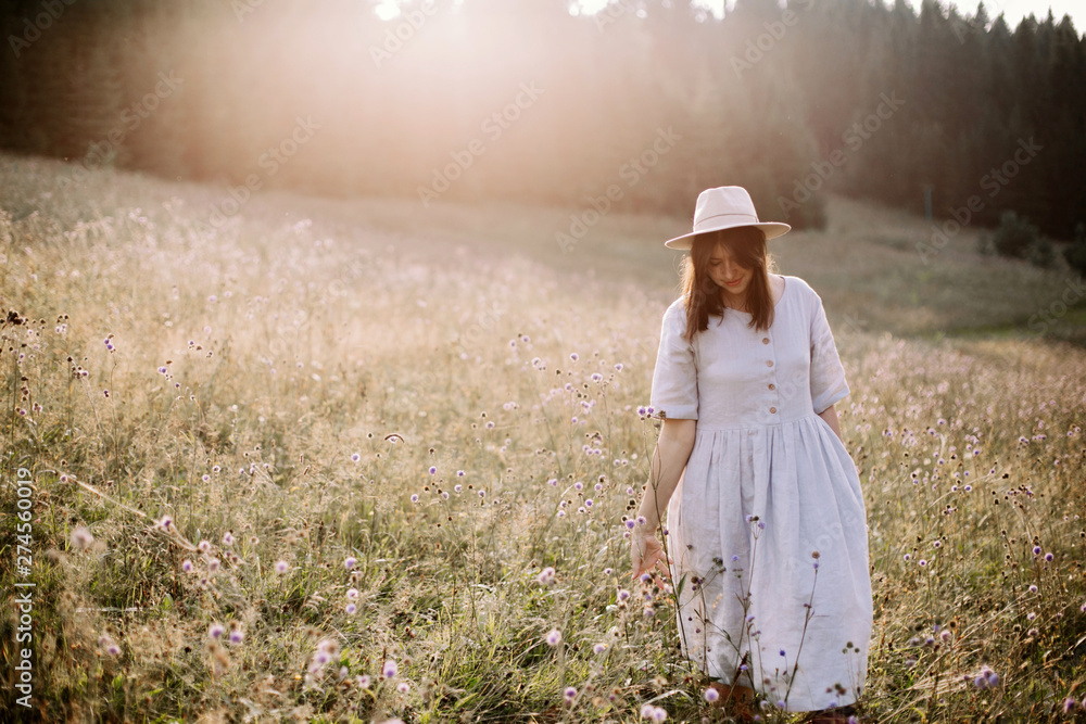 Stylish girl in rustic dress walking in wildflowers in sunny meadow in mountains. Boho woman relaxing in countryside flowers at sunset, rural simple life. Atmospheric image. Space text