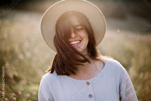 Stylish girl in rustic dress smiling and waving hair in sunny meadow in mountains. Portrait of happy boho woman in countryside at sunset, positive vibes, rural simple life. Atmospheric image