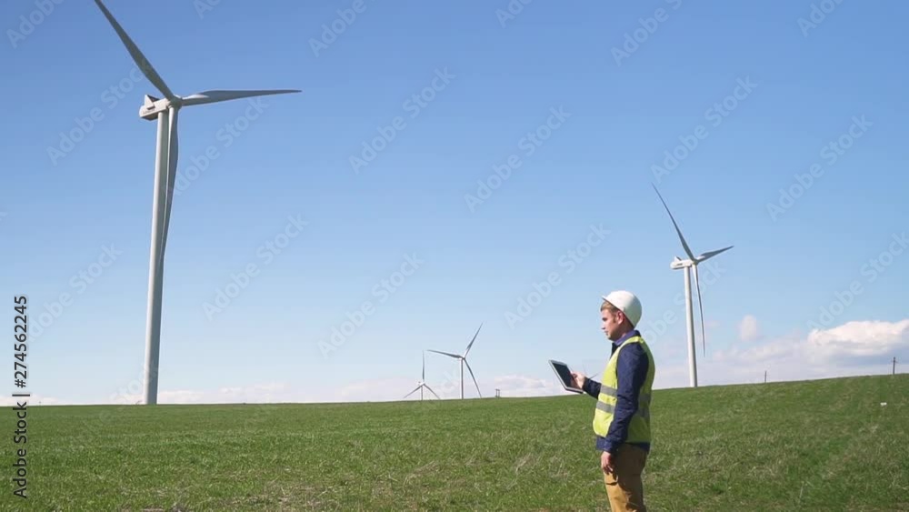 Adult or mature specialist man in protective uniform wear standing with portable tablet in hands against landscape wind turbine power station on background. Male checking information on device Adult or mature specialist man in protective uniform wear standing with portable tablet in hands against landscape wind turbine power station on background. Male checking information on device