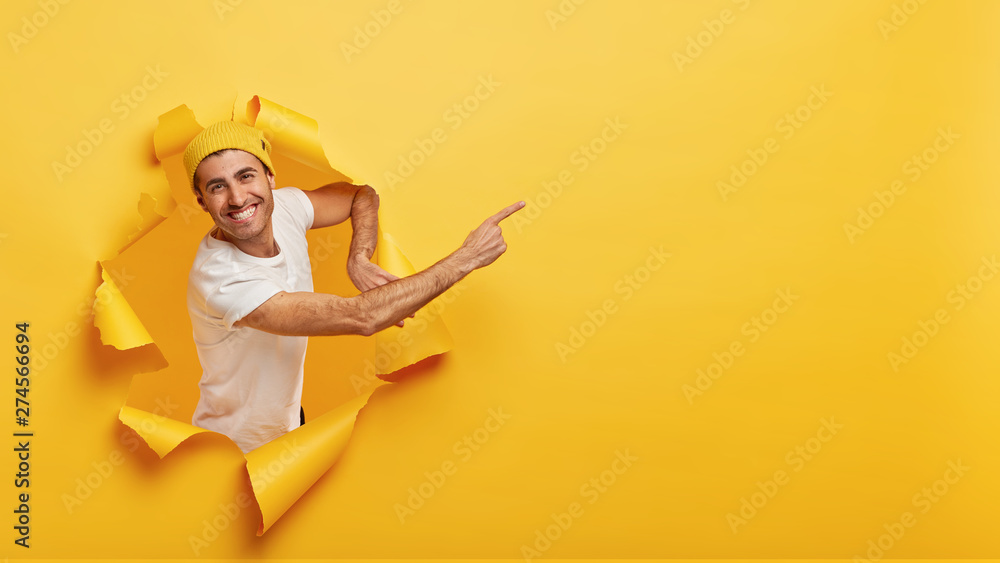 Smiling young handsome man stands in paper hole, points at free space, advertises or shows way, wears white t shirt, suggests going there, yellow background, has nice offer or gives opportunity
