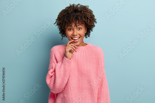 Curious happy woman keeps index finger on lips  looks with pleased facial expression  listens fine latest news from friend  enjoys wonderful time  dressed in loose sweater  has good feelings