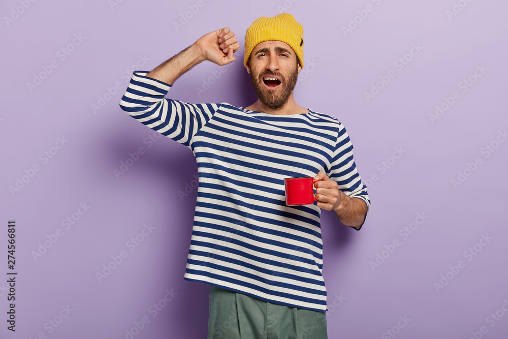 Morning starts with coffee. Sleepy man stretches after waking up, drinks hot beverage to feel refreshed, holds red cup, says good morning, wears casual clothes. People, awakening, drinking concept