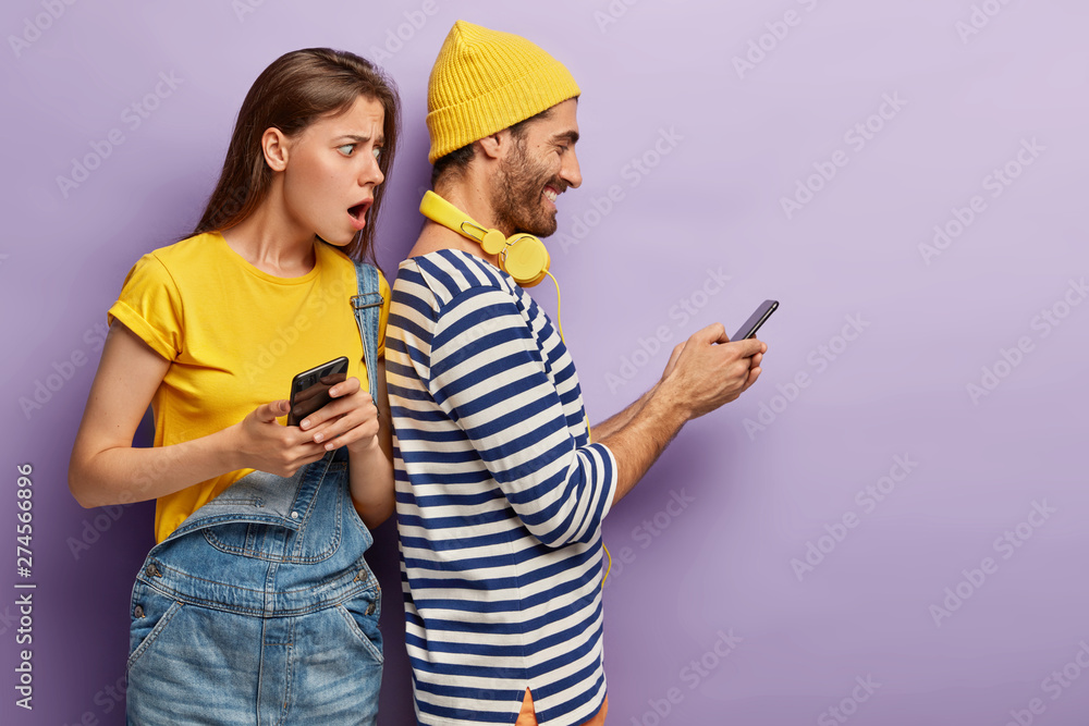 Suspicious jealous puzzled wife wants to spy sms message on husbands device, suspects of betrayal, looks over back, has embarrassed gaze. Young couple use modern gadgets for online communication - obrazy, fototapety, plakaty 
