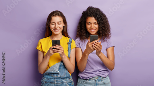 Two mixed race beautiful women send text messages on mobile phones, being addicted to modern technologies, make shopping online, have glad facial expressions, stand next to each other on purple wall photo