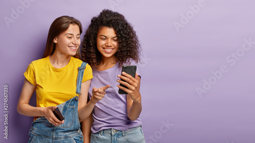 Pleased female friends watch something interesting on mobile phone, have happy relaxed expressions, focused in device, enjoy using modern technologies, isolated over purple wall with empty space photo