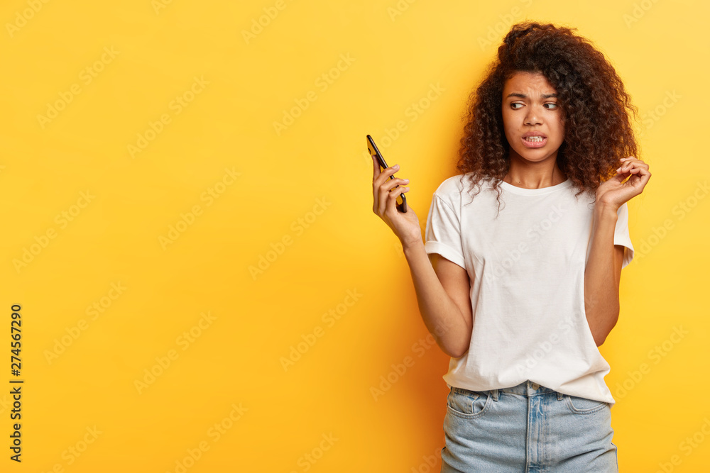 Beautiful female with frizzy Afro hairstyle, looks in displeasure at cell phone, receives call from former boyfriend, holds mobile phone on distance, ignores communication, afraids of something Beautiful female with frizzy Afro hairstyle, looks in displeasure at cell phone, receives call from former boyfriend, holds mobile phone on distance, ignores communication, afraids of something
