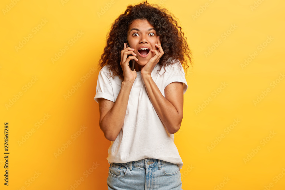 Sociable friendly impressed curly African American teenage girl holds breath from awesome news, calls friend via mobile phone, keeps mouth widely opened, dressed in casual white t shirt and jeans. Sociable friendly impressed curly African American teenage girl holds breath from awesome news, calls friend via mobile phone, keeps mouth widely opened, dressed in casual white t shirt and jeans.