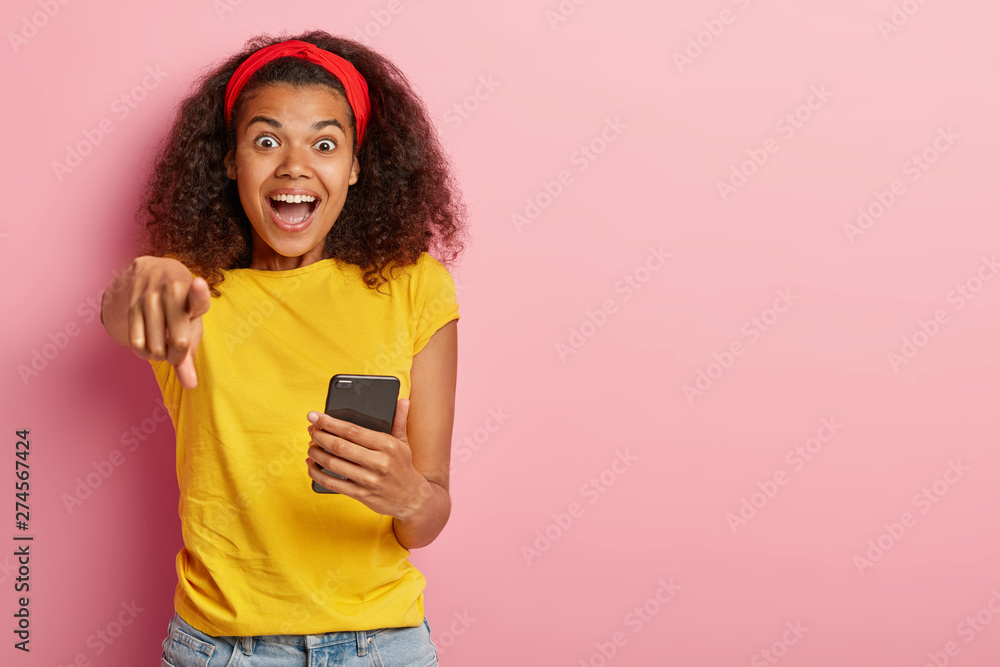 Emotional beautiful Afro female points at camera with wonder, expresses happiness, holds smartphone device, enjoys virtual communication, checks mail, uses free wifi wears yellow casual t shirt Emotional beautiful Afro female points at camera with wonder, expresses happiness, holds smartphone device, enjoys virtual communication, checks mail, uses free wifi wears yellow casual t shirt