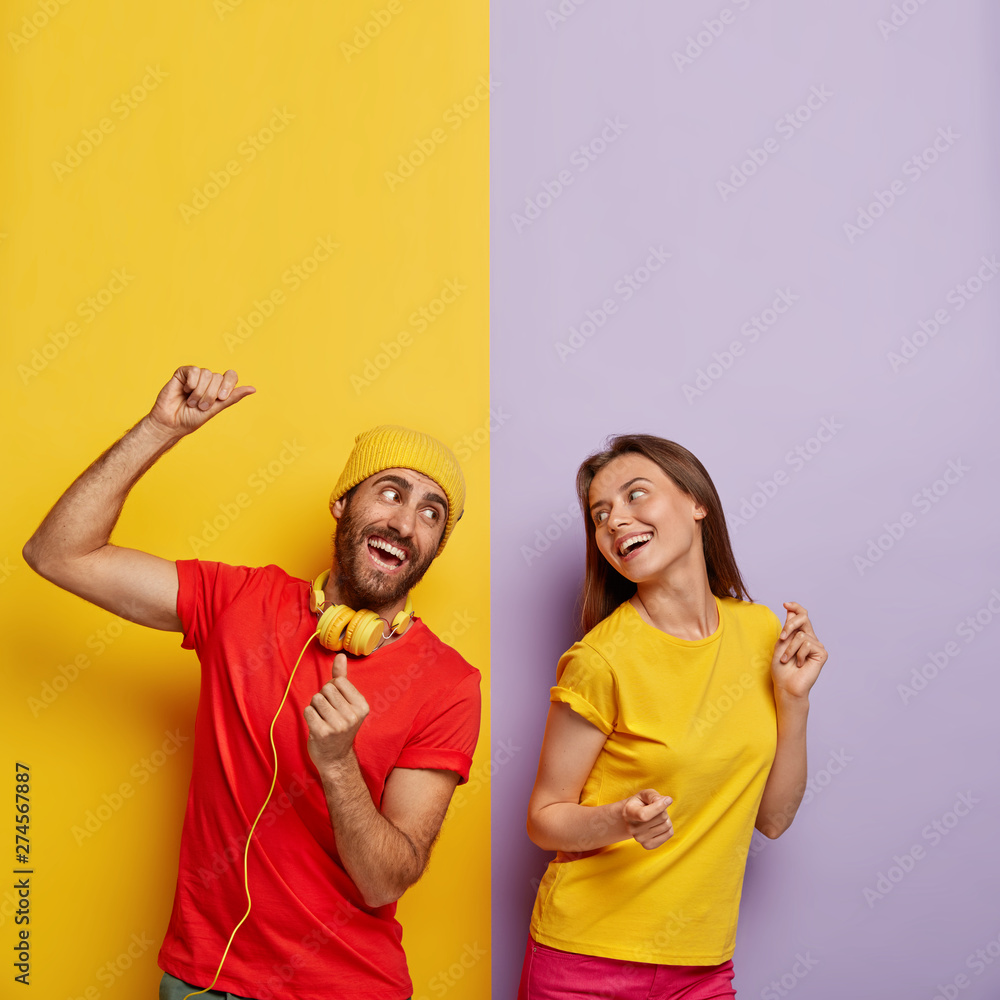 Positive couple listen music together, dance and move actively, being in good mood. Cheerful man in red t shirt, headphones on neck, spends free time with girlfriend. People and entertainment concept - obrazy, fototapety, plakaty 