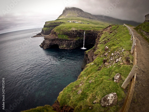 Wide extra panoramic view on Faroe Island famous waterfall with small town above it. Cloudy sky and green hills in the background.