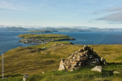 View on multiple islands with small city down the hill. Green grass, blue ocean and cloudy sky in the background. Stone pile placed for orientation purpose in front. Faroe Islands.