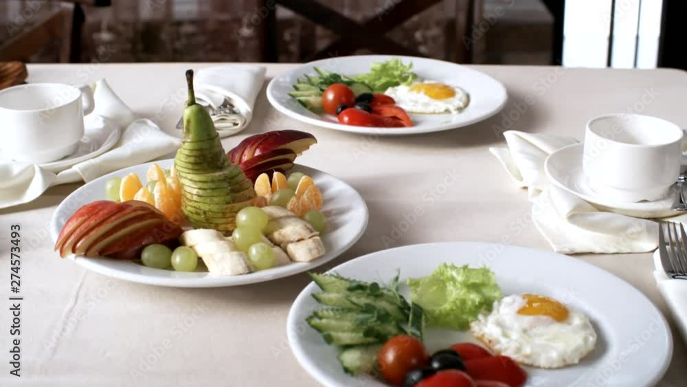 Close-up panning object shot of table in hotel restaurant set for healthy breakfast for two, with fried egg, fresh vegetables, fruit platter, toast and jam, with cutlery and empty cups beside plates Close-up panning object shot of table in hotel restaurant set for healthy breakfast for two, with fried egg, fresh vegetables, fruit platter, toast and jam, with cutlery and empty cups beside plates