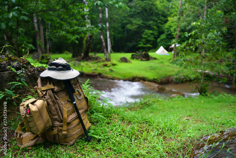 backpack with medicine bag and hiking or walking pole with hat for trekking and camping tent on stream with tree in green grass jungle for holiday relax and vacation travel trip with adventure nature