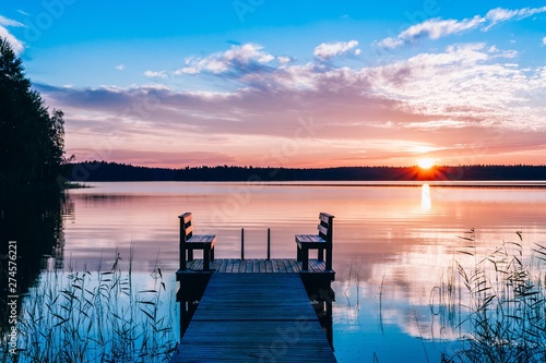 Idyllic view of the long pier with wooden bench on the lake. Sunset or sunrise over the water. © nblxer