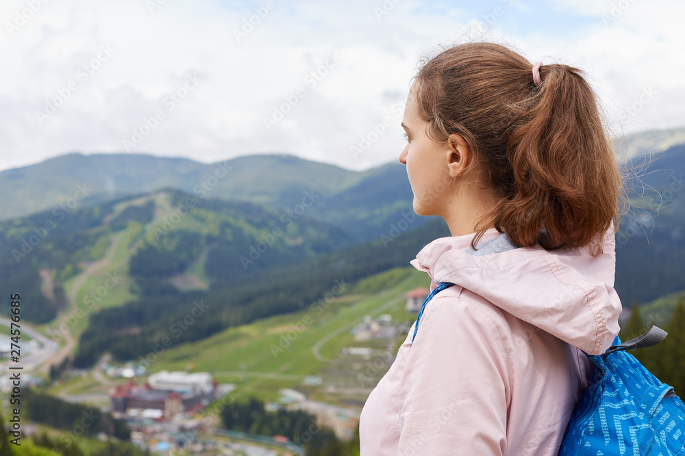 Obraz premium Profile of experienced traveller with pony tail wearing backpack and rose jacket, enjoying mountain landscape, having travelling mood, being focused on trip route. People and active lifestyle concept.