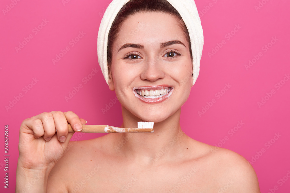 Close up portrait of attractive caucasian smiling dark haired woman isolated on rose studio, posing with towel on head and bare shoulders, has perfect skin, looks at camera with tooth brush in hand. Close up portrait of attractive caucasian smiling dark haired woman isolated on rose studio, posing with towel on head and bare shoulders, has perfect skin, looks at camera with tooth brush in hand.