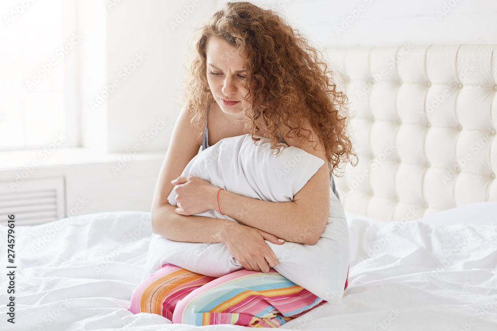 Indoor shot of young emotional lady feeling sharp pain, hugging her pillow, pressing it tight to her body, being ill, having unpleasant facial expression, wearing pyjamas. Health care concept. Indoor shot of young emotional lady feeling sharp pain, hugging her pillow, pressing it tight to her body, being ill, having unpleasant facial expression, wearing pyjamas. Health care concept.