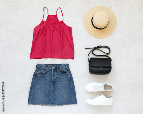 Pink cami top, blue denim mini skirt, straw boater hat, small black cross body bag, white sneakers on grey background. Overhead view of woman's casual outfits. Flat lay, top view. Trendy summer look.