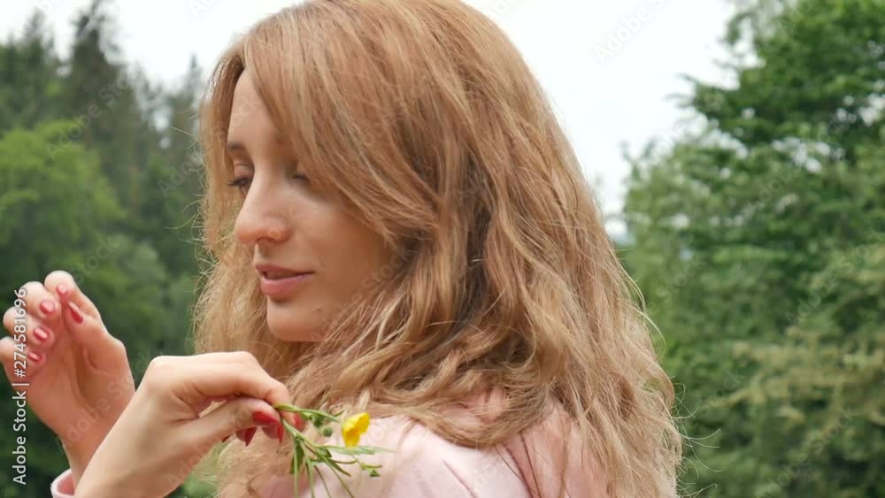 Beautiful curly red-haired woman holding a yellow wild flower. Portrait against the forest and mountains. Summer vacation, spring freshness, mother's day, allergy free Beautiful curly red-haired woman holding a yellow wild flower. Portrait against the forest and mountains. Summer vacation, spring freshness, mother's day, allergy free
