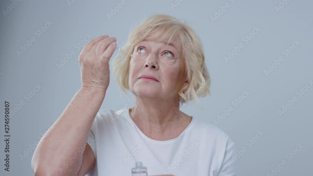 Blonde older woman putting fake tears on her clean face pretending to cry isolated on grey background. Blonde older woman putting fake tears on her clean face pretending to cry isolated on grey background.